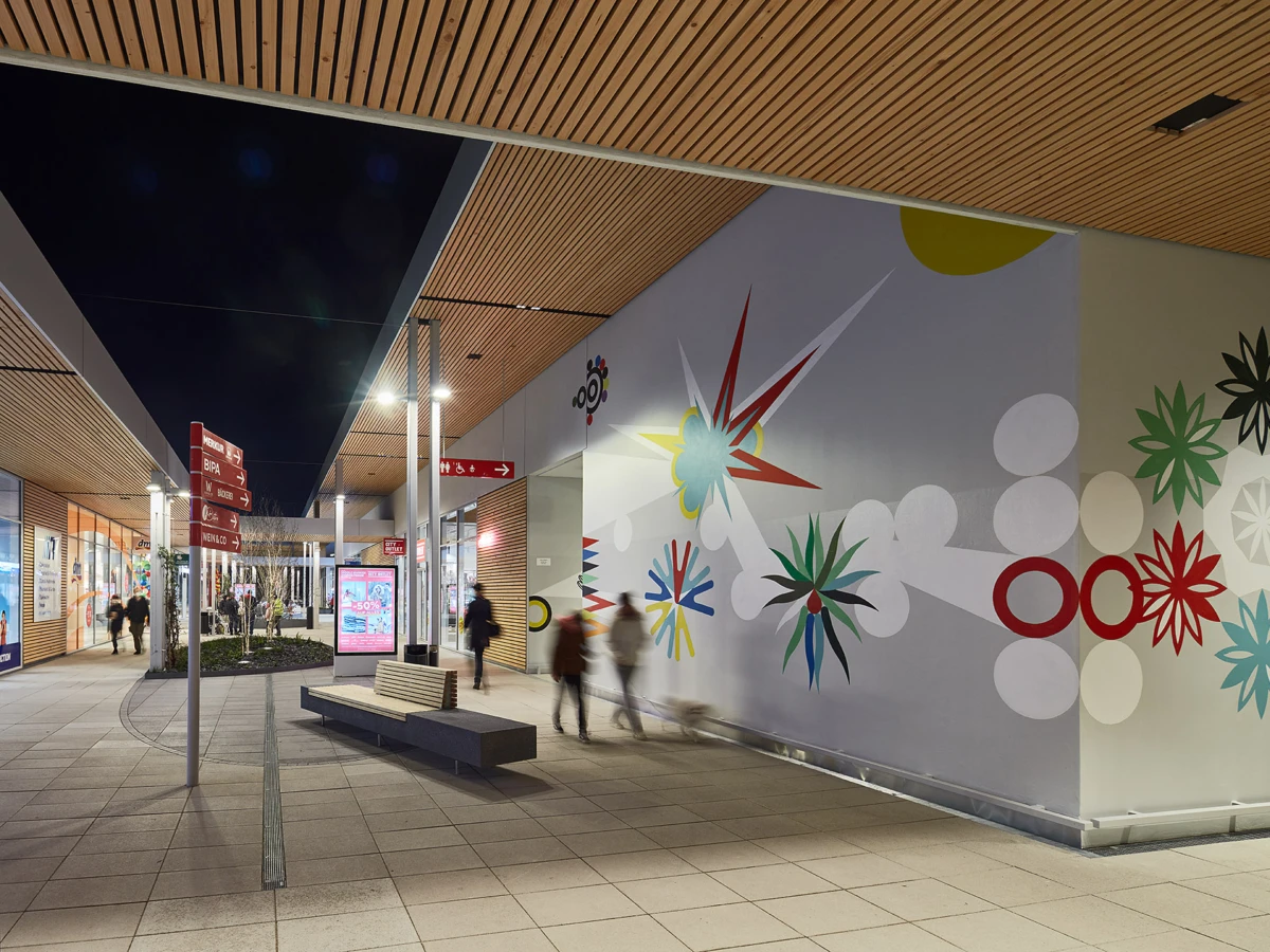 Nighttime covered pedestrian promenade with a colorful mural on the right wall, wooden ceiling panels, benches, and pedestrians.