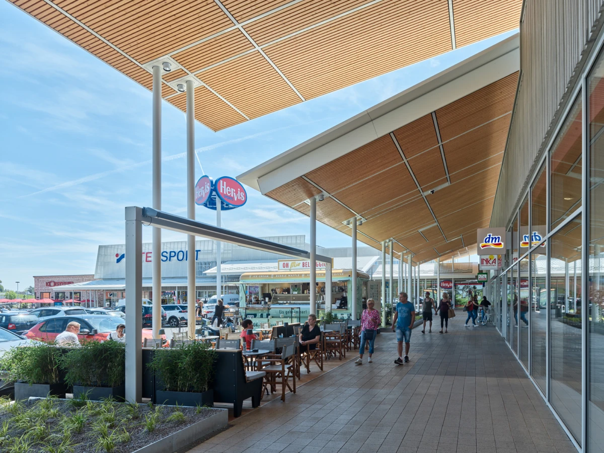Outdoor shopping promenade under a wooden canopy with cafe seating; shoppers stroll beside glass storefronts.