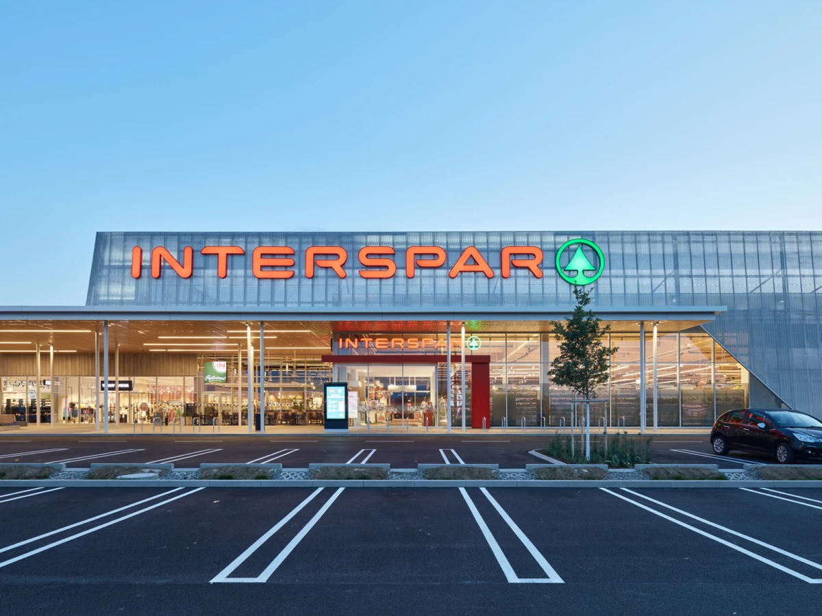 Front view of an INTERSPAR supermarket storefront with orange logo and green circular emblem, glass façade, and empty parking lot.