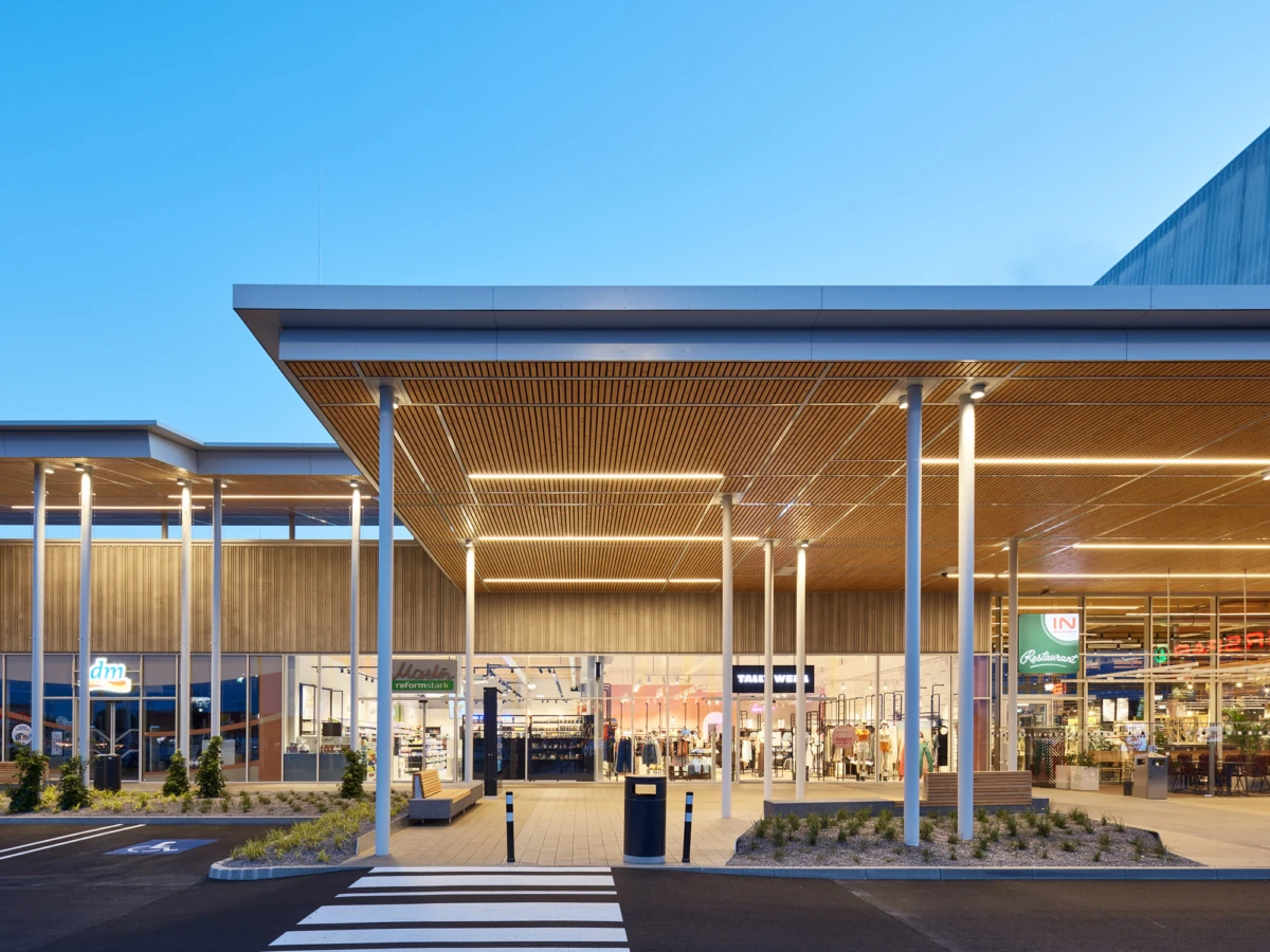 Shopping center entrance under a long wooden slat canopy, glass storefronts, and shoppers inside; well-lit, modern retail plaza.