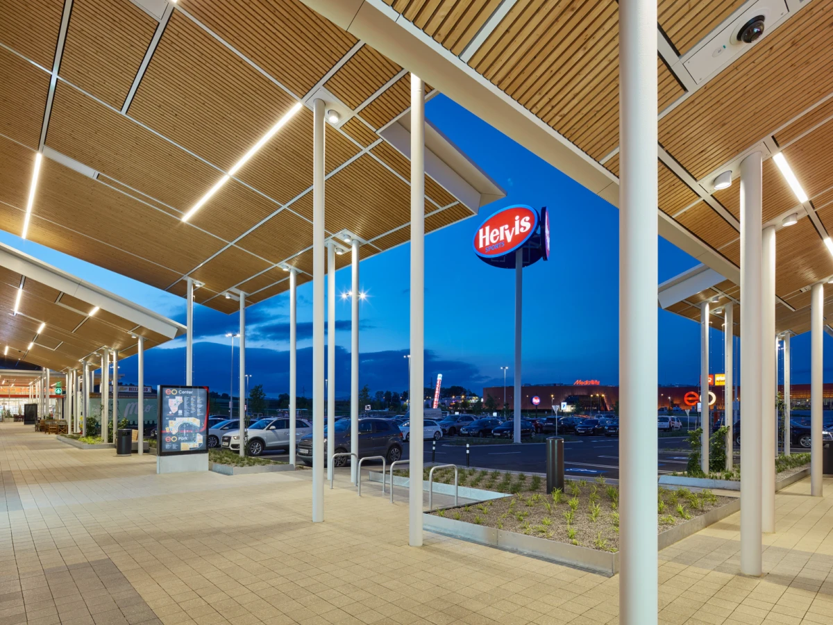 Empty covered walkway of a shopping plaza at dusk, white columns, wooden ceiling panels, and a parking lot in the background.