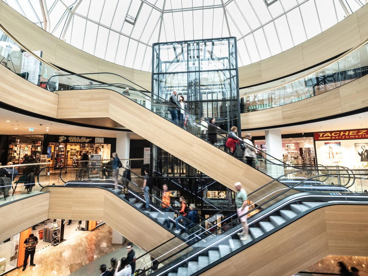 Three-story shopping mall atrium with curved wooden escalators circling a glass elevator shaft; shoppers ride escalators and browse storefronts.
