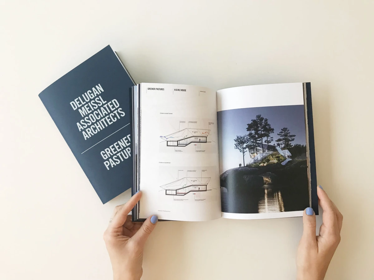 Hands hold open architecture book; left page diagrams, right page lakeside forest house; blue cover reads Delugan Meissl Associated Architects.