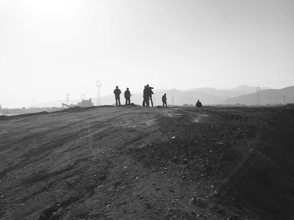 Group of silhouetted people standing on a dirt mound in a desolate landscape with hills and power lines in the distance.