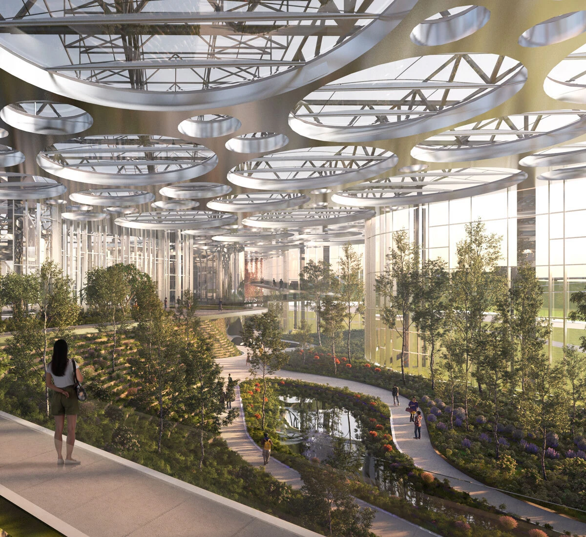 Modern glass-walled indoor garden atrium with circular ceiling rings; visitors walk along an elevated path above a water garden.