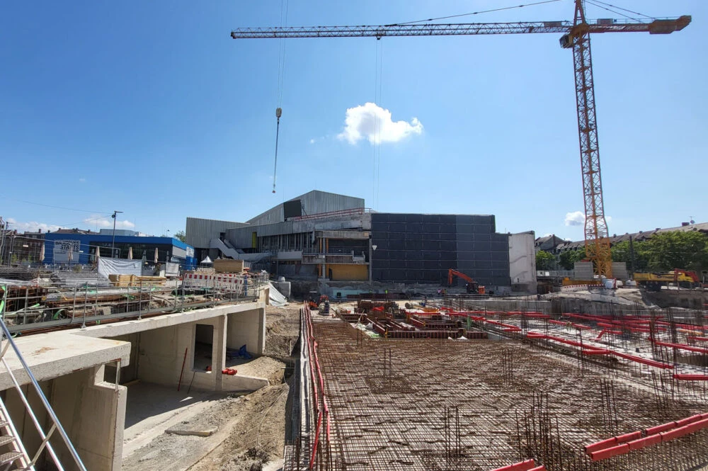 Tower crane over a large construction site with rebar grid, concrete structures, and a hoist hook against a clear blue sky.