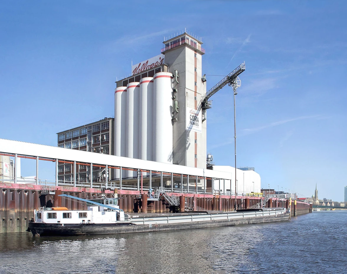Industrial riverfront with tall white grain silos, a crane, and a docked barge under a blue sky.