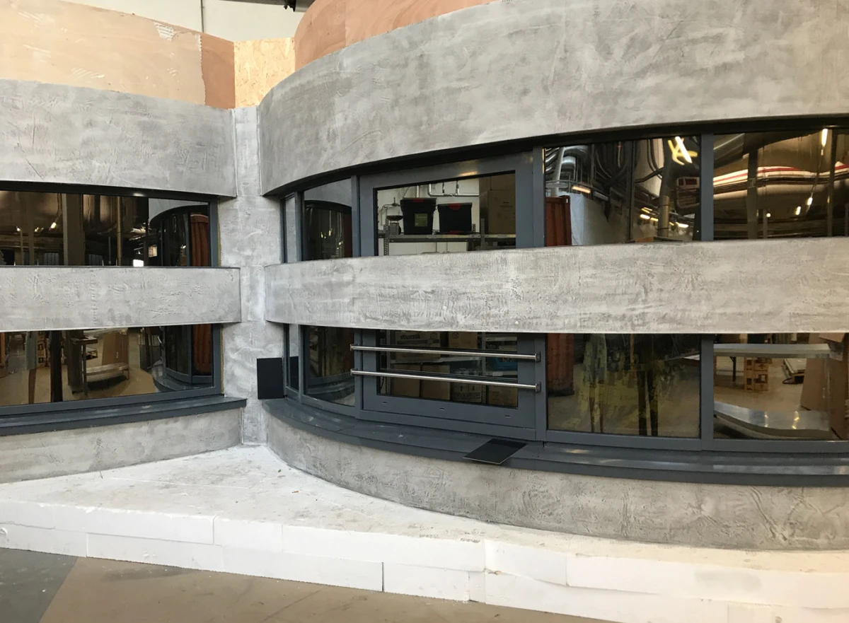 Curved concrete wall with large glass windows and a metal railing; interior workshop visible through the panes.