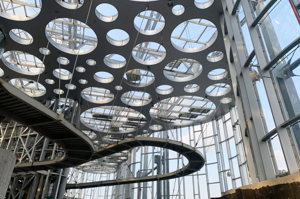 Spiral steel pedestrian ramp descends through a glass atrium with circular skylight panels.