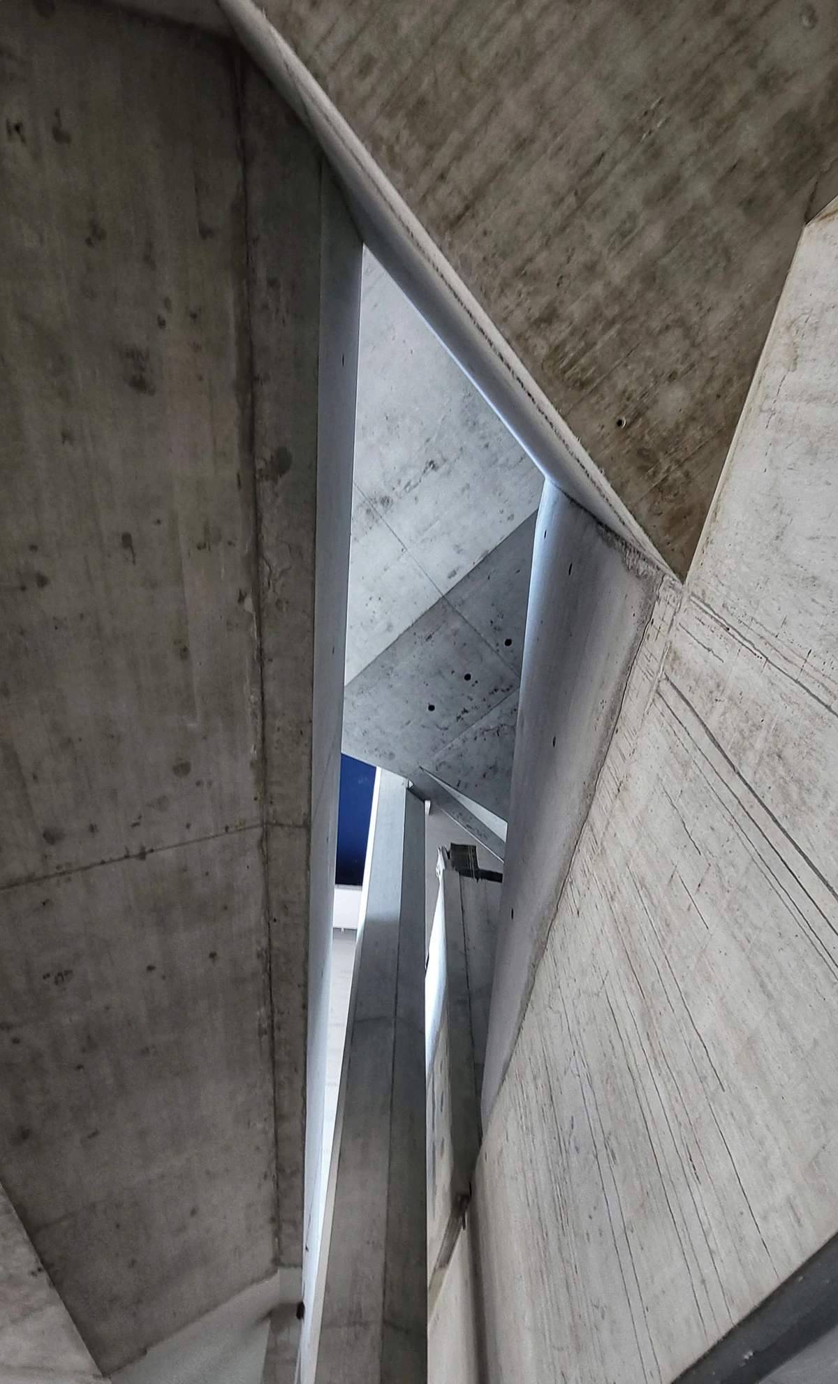 Narrow interior concrete stairwell with angled slabs and vertical supports; blue sky visible through the opening.