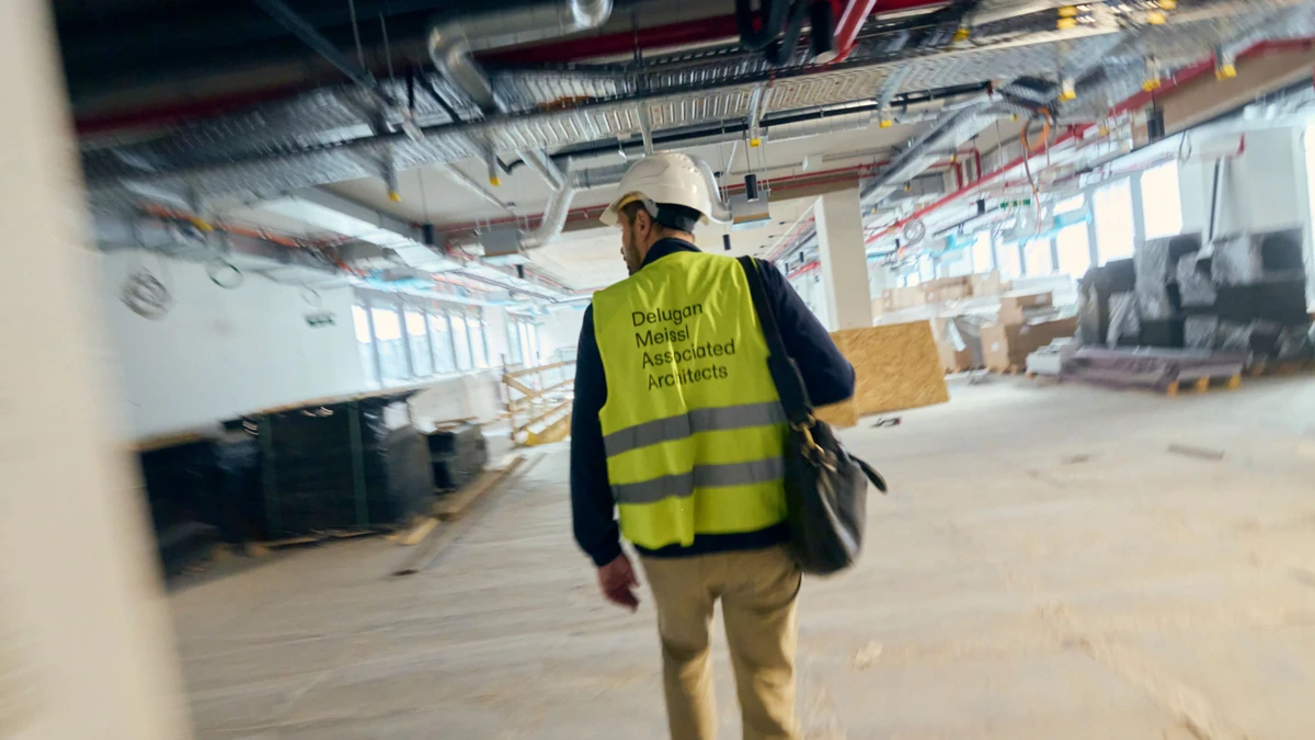 Construction worker in high-visibility safety vest and white hard hat walks through a partially finished interior with exposed pipes.