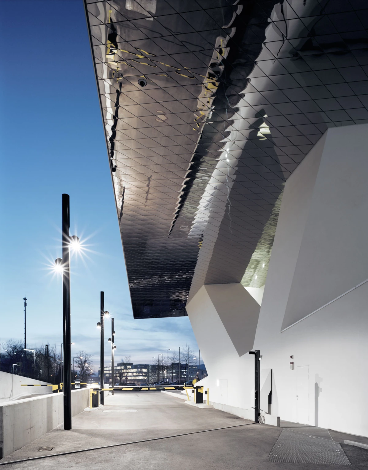 Silver metallic canopy over a white angular building, with tall street lamps along a concrete driveway and a distant barrier gate.
