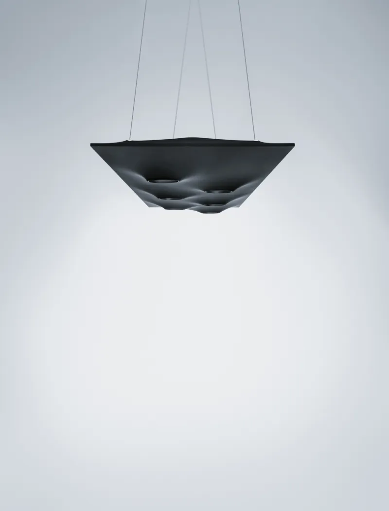 Black square pendant lamp with circular recesses, suspended by thin cables in a minimal white studio backdrop.
