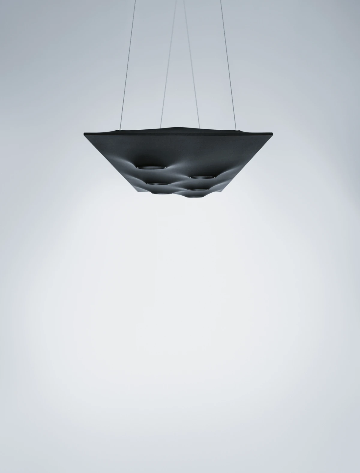 Black square pendant lamp with circular recesses, suspended by thin cables in a minimal white studio backdrop.